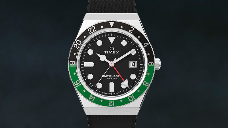 Front-facing product shot of the Q Timex GMT watch with a black dial, bold luminous indices, and a red GMT hand. The bezel features a black-and-green split with 24-hour markings, and the date window is positioned at 3 o'clock. The black strap and sharp case lines reflect a sporty yet refined aesthetic.