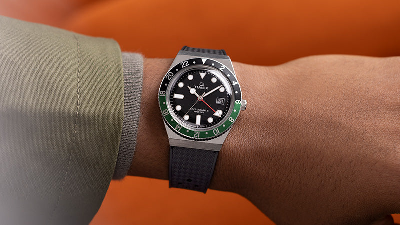 Wrist shot of the Q Timex GMT watch worn on the left wrist, displaying the timepiece against an orange backdrop. The black dial, luminous markers, and dual-tone bezel in green and black stand out prominently. The watch is paired with a textured black strap, creating a bold and modern look.