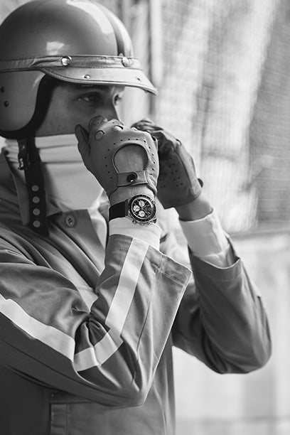 Black-and-white photo of a motorsport driver wearing vintage racing gear, including a helmet and gloves. The driver adjusts his helmet strap, while a bold chronograph Q Timex watch is visible on his wrist, suggesting a blend of timing precision and classic racing heritage.