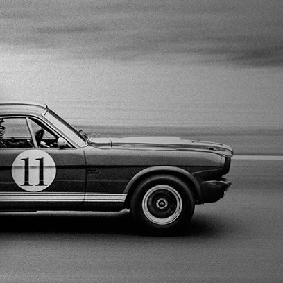 Monochrome image of a vintage racing car with the number 11 on the door, speeding along a blurred racetrack. The shot captures motion and energy, evoking the golden era of motorsports, aligning with the Q Timex racing chronograph theme.