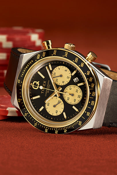 Close-up product shot of a Q Timex chronograph watch resting on a red poker table. The watch features a black and gold tachymeter bezel, gold subdials, and pushers, paired with a dark leather strap. A stack of poker chips in the background hints at a theme of high-stakes style and sophistication.