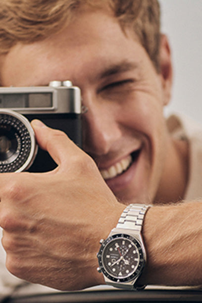 Portrait of a smiling man holding a vintage camera up to his face. He wears a silver Q Timex chronograph watch with a black dial and subdials, complementing the nostalgic theme. The relaxed, candid moment reflects timeless style and a love for classic craftsmanship.