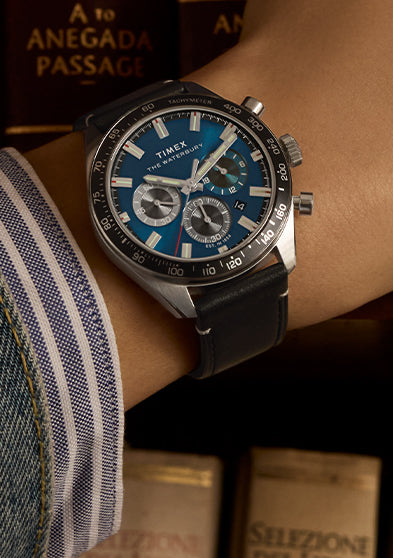 Close-up of the Timex Waterbury Chronograph on a wrist, featuring a striking blue dial, three subdials, and a tachymeter bezel. The watch is paired with a black leather strap and worn with a denim jacket and striped shirt cuff. In the background, leather-bound books—one titled “A to Anegada Passage”—add a refined, intellectual atmosphere.