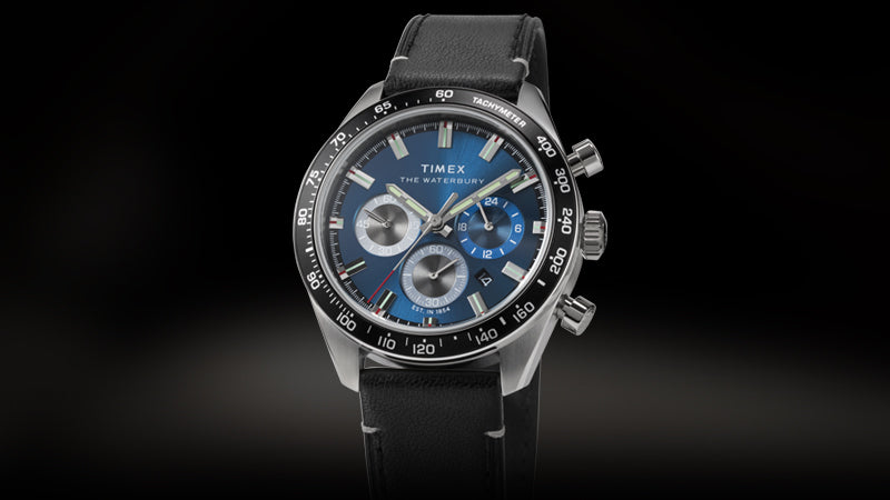 Front-facing product image of the Timex Waterbury Chronograph featuring a vibrant blue dial with silver-tone subdials, applied markers, and tachymeter bezel. The watch is paired with a black leather strap, and showcased against a clean, dark background for a dramatic presentation.