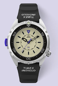 A front-facing product image of the Timex x Dimepiece watch featuring a cream dial, black unidirectional bezel, bold INDIGLO® branding on the strap, and a purple crown. 