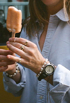 A candid lifestyle image showing a woman wearing the Timex x Dimepiece watch while holding a popsicle. The watch is styled with gold and pearl jewelry, adding contrast to the utilitarian design of the timepiece.