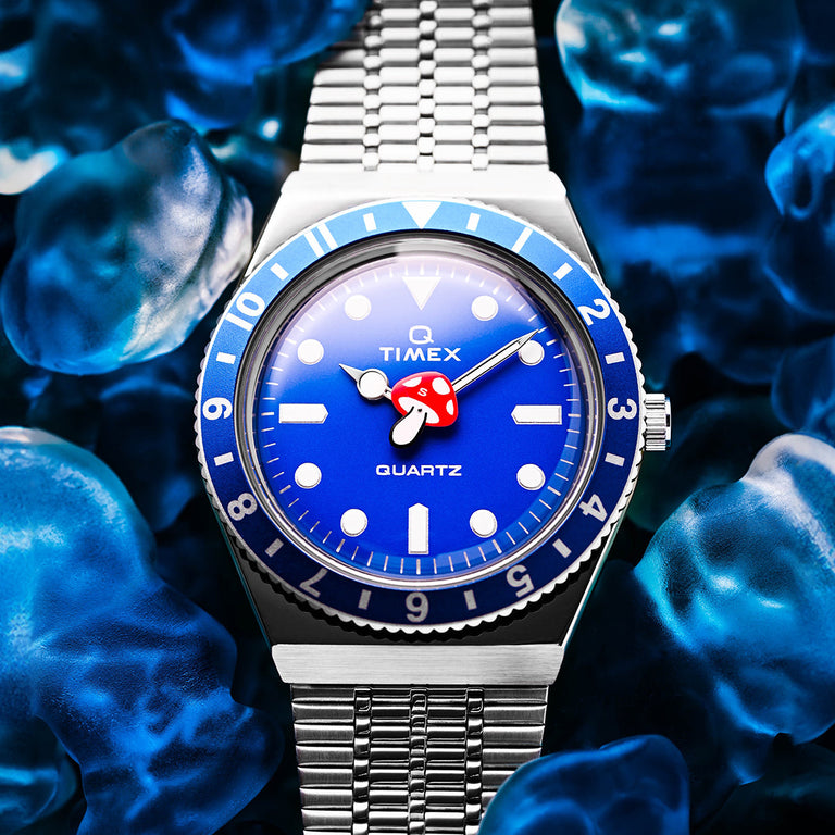 A Timex x seconde/seconde/ Q Timex watch with a vibrant blue dial, blue-and-silver rotating bezel, and stainless steel bracelet. The dial features playful design details, including a red mushroom-shaped seconds hand, set against a backdrop of translucent blue objects for a bold, artistic presentation.