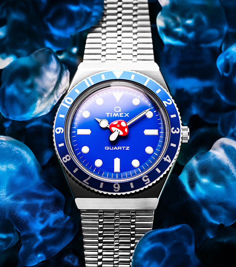 Timex x seconde/seconde/ Prequel Edition watch with a vibrant blue dial and bezel, set against a background of translucent blue gummy bears. The dial features a whimsical red-and-white mushroom as the hour hand, and classic diver-style indices and hands.