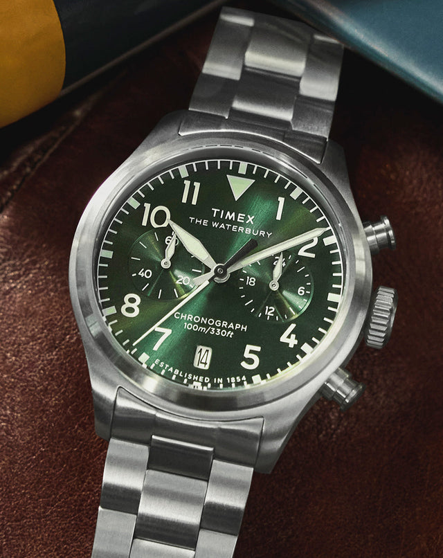 Timex Waterbury Ace Chronograph with a green dial and stainless steel bracelet resting on a brown leather surface, highlighting its brushed steel case and chronograph subdials.