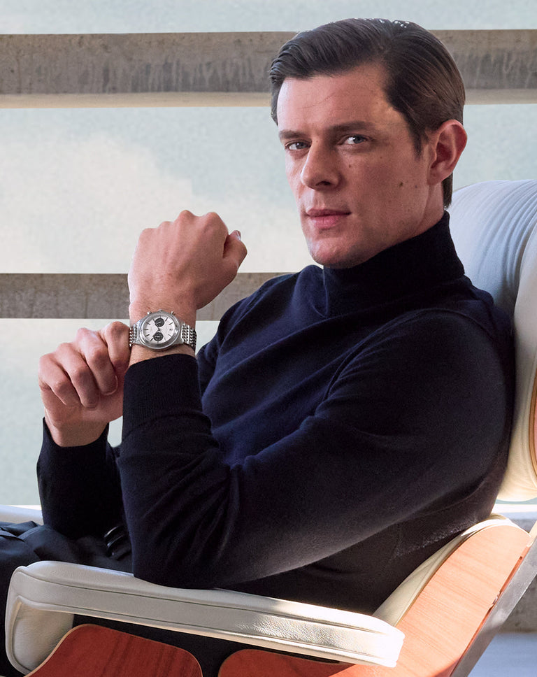 A man seated in a modern chair wearing a black turtleneck and silver wristwatch, looking confidently toward the camera with his left arm raised to display the watch.