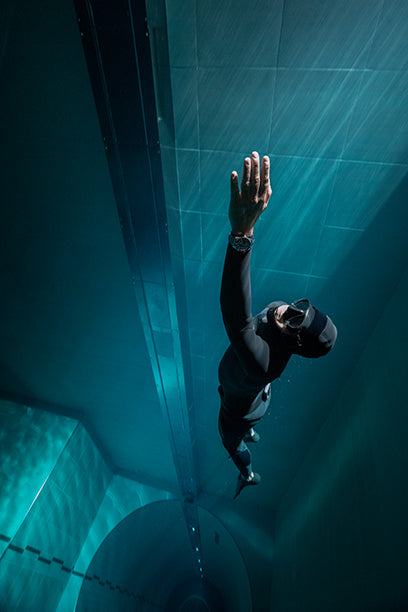 Freediver extends one arm upward while swimming in a deep vertical pool, wearing the Timex Deepwater Meridian 200.