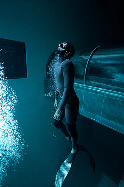 Freediver in a wetsuit exhales bubbles underwater near a tunnel opening, wearing the Timex Deepwater Meridian 200.