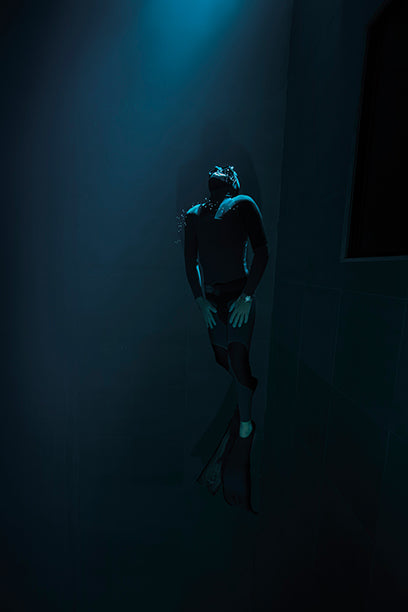 Freediver in a black wetsuit and fins ascends toward the surface in deep water, illuminated by a shaft of light from above.