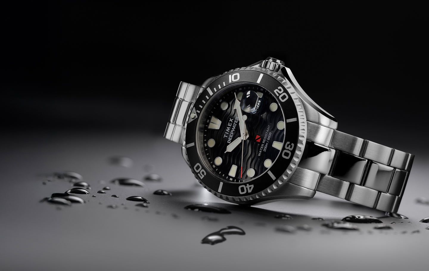 Timex Deepwater dive watch with stainless steel bracelet and black wave-textured dial, displayed at an angle with water droplets on a dark surface.