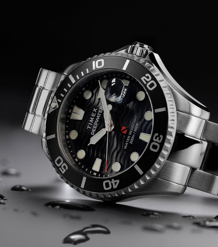 Timex Deepwater dive watch with stainless steel bracelet and black wave-textured dial, displayed at an angle with water droplets on a dark surface.