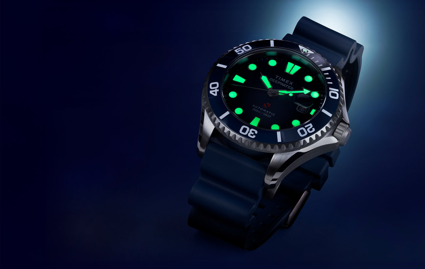 A Timex Deepwater automatic dive watch with a stainless steel case, black rotating bezel, and luminous green markers glowing in the dark, set against a deep blue background.
