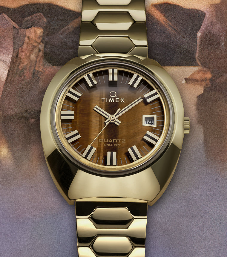 Timex Q wristwatch with a gold-tone case and bracelet, featuring a tiger’s eye-pattern dial, bold white hour markers, date window at 3 o’clock, and set against a soft, painted landscape background.