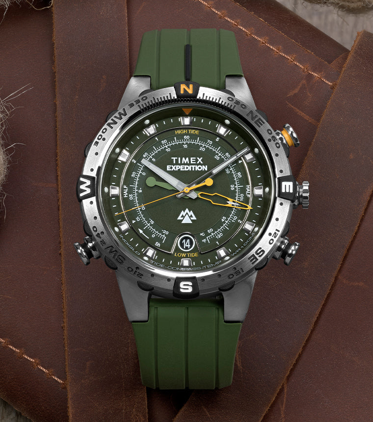Timex Expedition® Tide-Temp-Compass watch with green dial and strap, featuring a stainless steel case, rotating compass bezel, and multiple sub-dials, resting on a brown leather surface with a coiled rope.