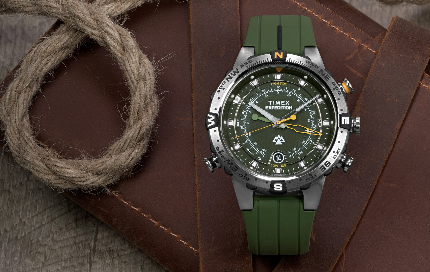 Timex Expedition® Tide-Temp-Compass watch with green dial and strap, featuring a stainless steel case, rotating compass bezel, and multiple sub-dials, resting on a brown leather surface with a coiled rope.