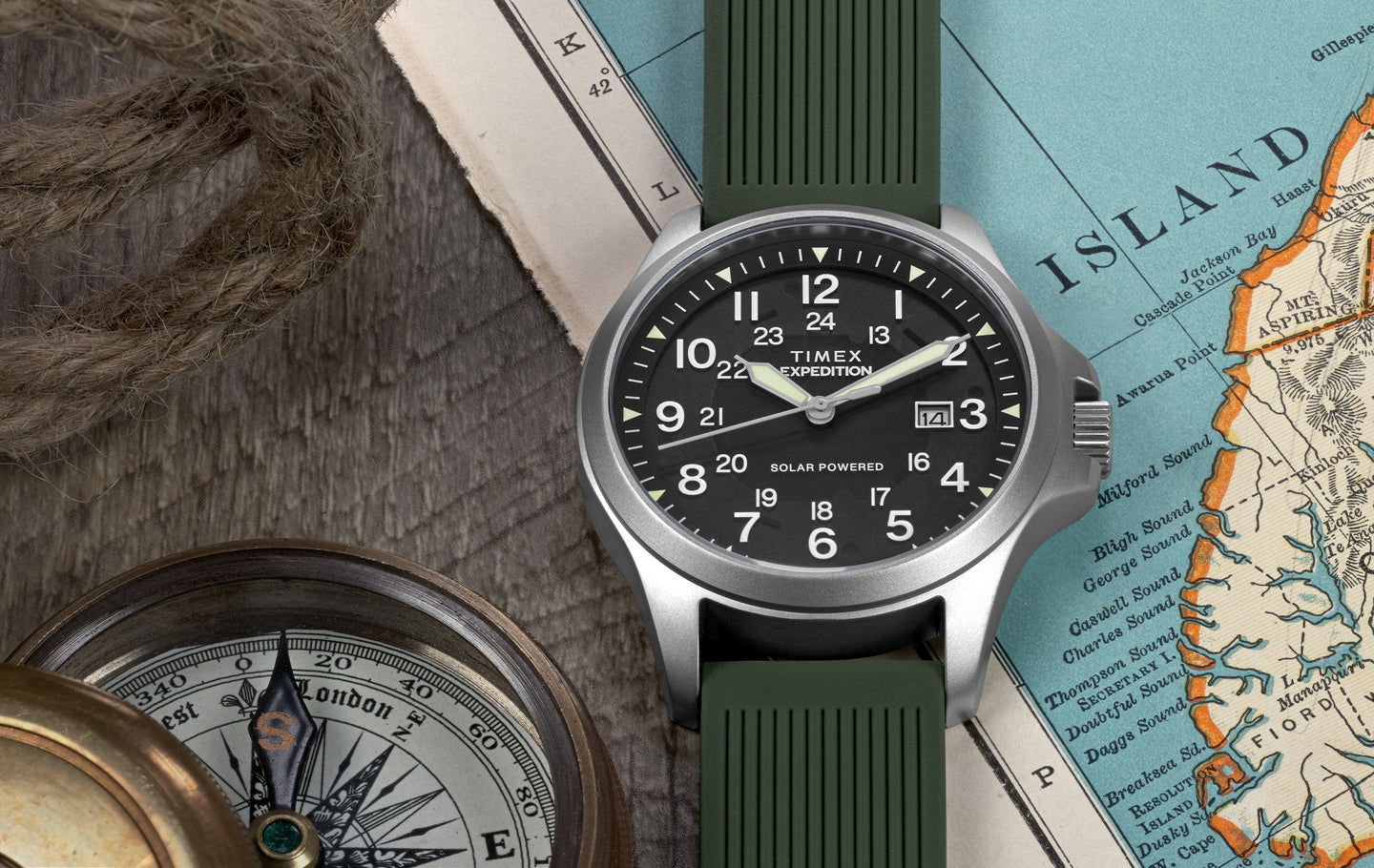 A Timex Expedition® Field Post Solar watch with a green silicone strap rests on a wooden surface beside a rope, a brass compass, and a vintage map. The watch has a black dial with white Arabic numerals, luminous hands, and a date window at 3 o’clock. The text on the dial reads “TIMEX Expedition” and “Solar Powered.”