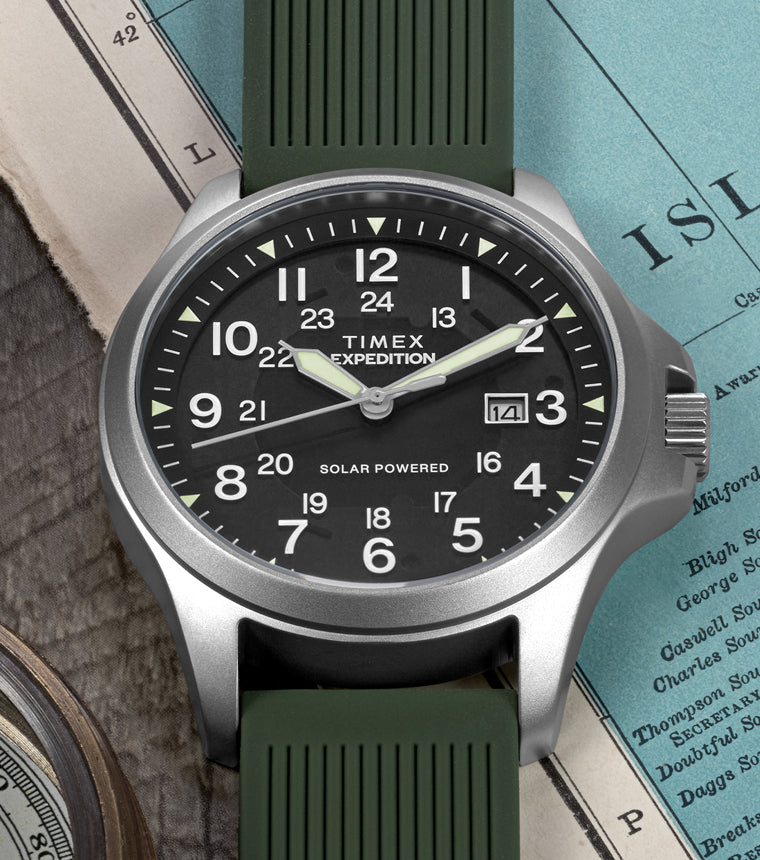 A Timex Expedition® Field Post Solar watch with a green silicone strap rests on a wooden surface beside a rope, a brass compass, and a vintage map. The watch has a black dial with white Arabic numerals, luminous hands, and a date window at 3 o’clock. The text on the dial reads “TIMEX Expedition” and “Solar Powered.”