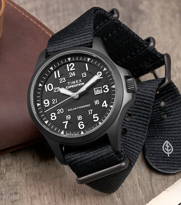 Timex Expedition® Field Post Solar black watch with recycled fabric strap, black dial, date display, and solar-powered movement on a leather journal.