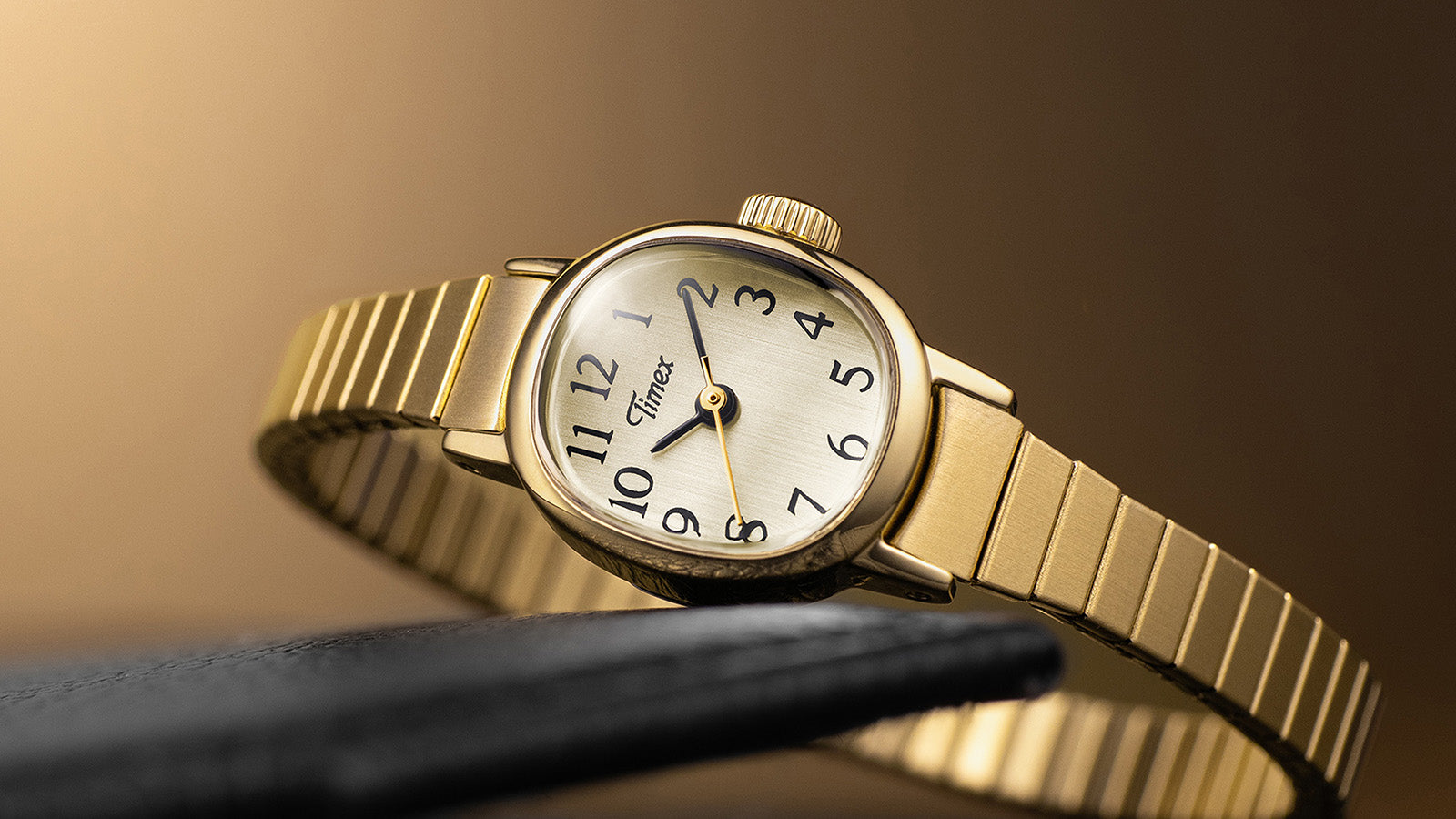 Timex Cavatina Mini gold-tone watch with expansion bracelet, resting on a black leather surface with a warm golden background.