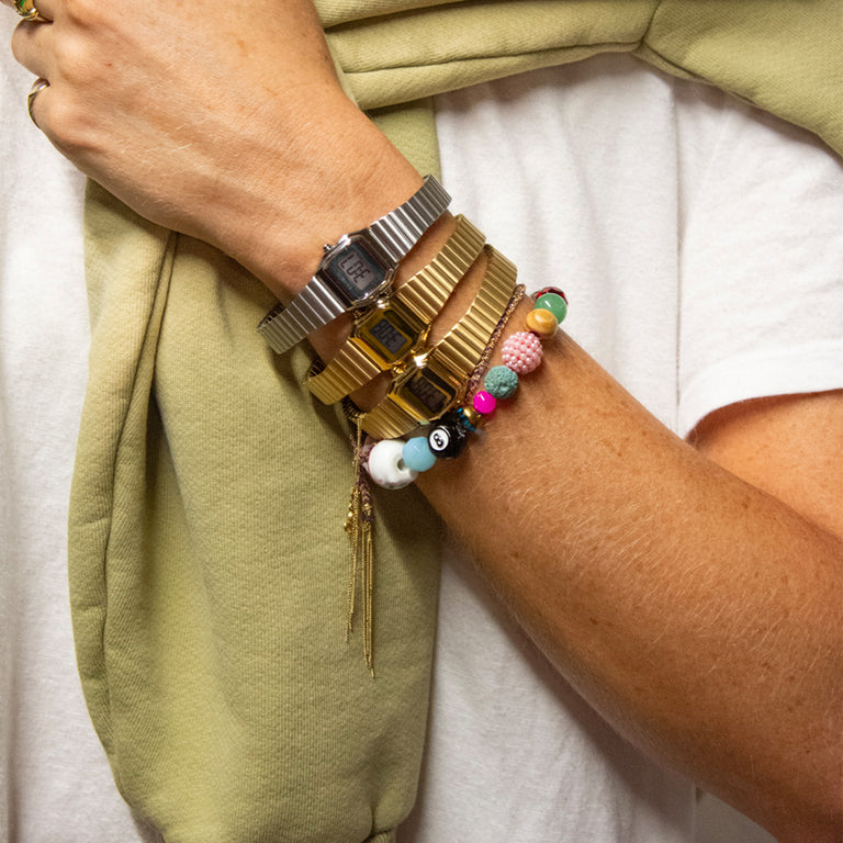 Close-up of a person wearing multiple Timex Teeny Tiny digital watches in gold and silver tones, stacked with colorful beaded bracelets and a light green sweatshirt draped over their shoulders.