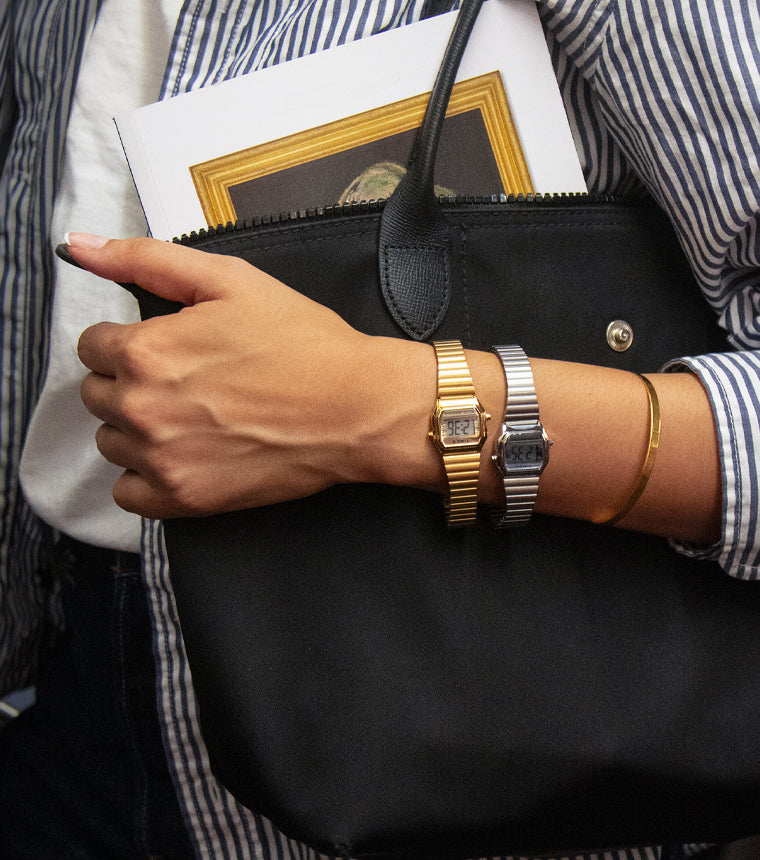 A person wearing two small rectangular Timex digital watches — one gold and one silver — on their wrist, along with a thin gold bracelet. They are holding a black handbag and a small framed print, dressed in a blue-and-white striped shirt over a white top.