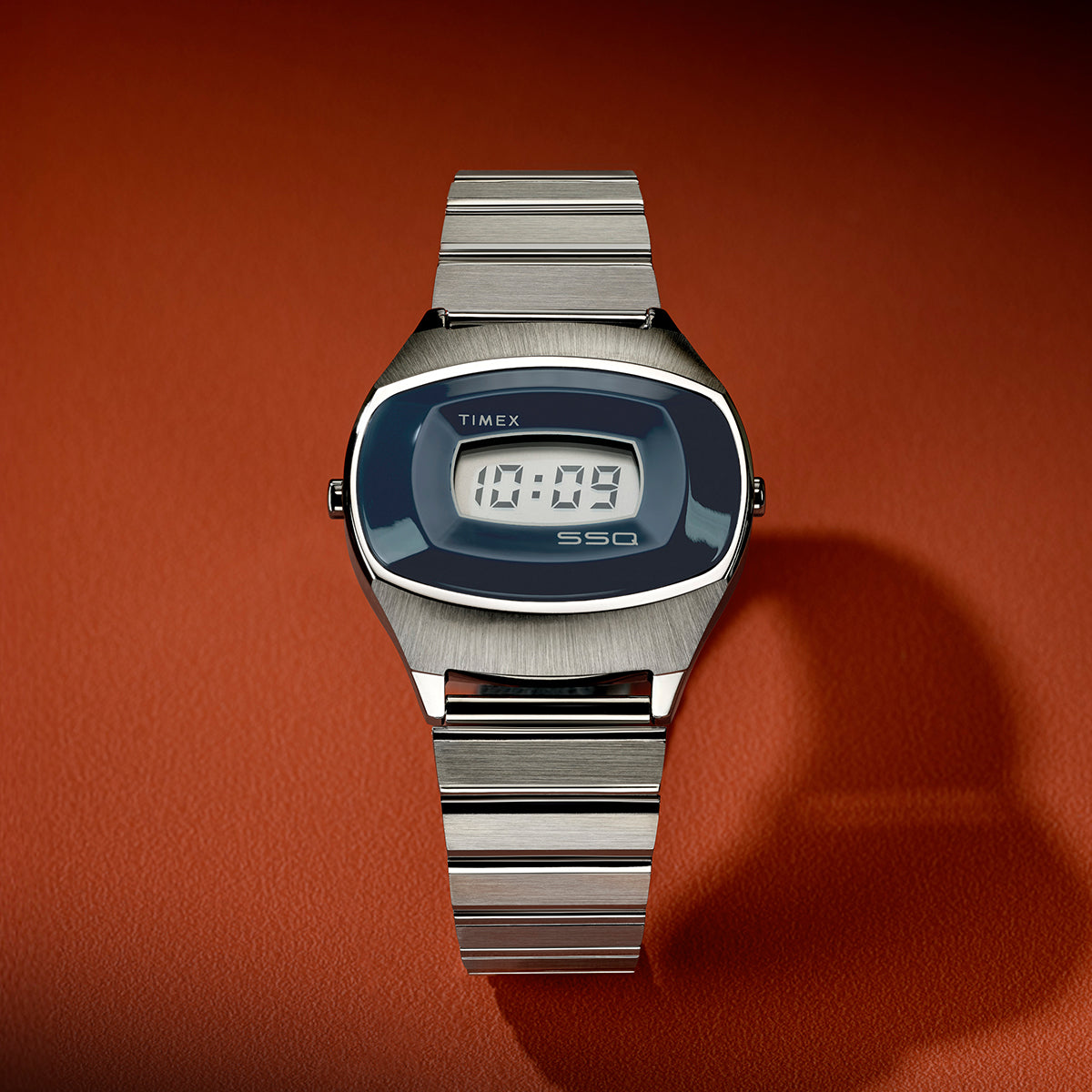 Front view of the Q Timex® 1975 SSQ Digital Reissue with brushed stainless-steel bracelet, blue digital display, and retro-inspired oval case on a warm orange leather surface.