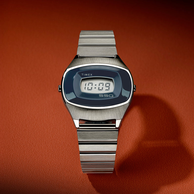 Front view of the Q Timex® 1975 SSQ Digital Reissue with brushed stainless-steel bracelet, blue digital display, and retro-inspired oval case on a warm orange leather surface.