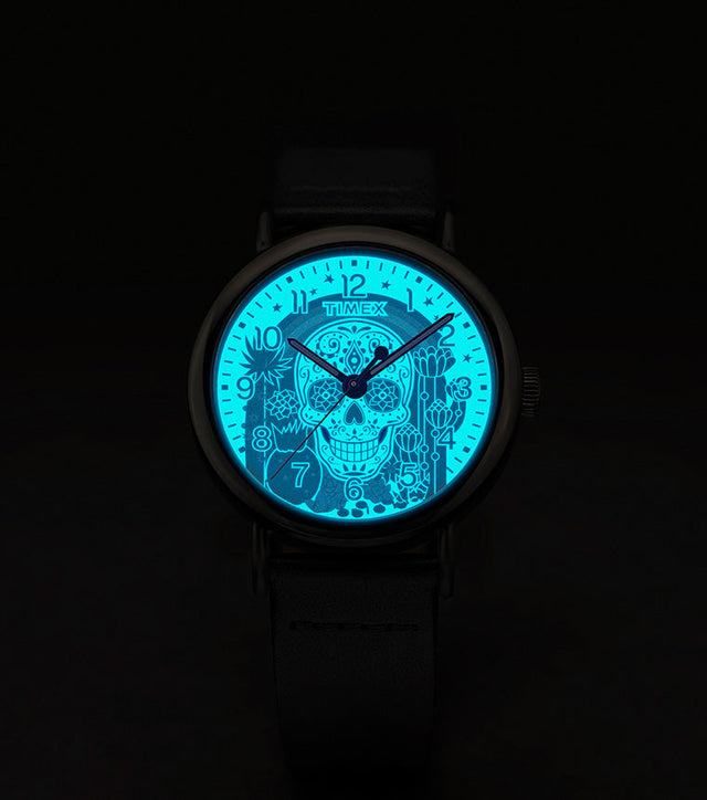 Timex Weekender Day of the Dead watch dial glowing in the dark with INDIGLO® backlight, revealing a bright blue sugar skull and floral details.