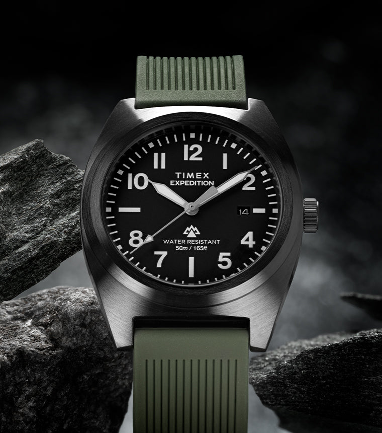 A Timex Expedition® Capstone watch with a black dial, white numerals, and luminous hands, displayed among rugged gray rocks. The watch has a brushed stainless steel case, olive green textured strap, and is labeled “Water Resistant 50m / 165ft.” The dark, moody lighting emphasizes its durable, outdoor-ready design.