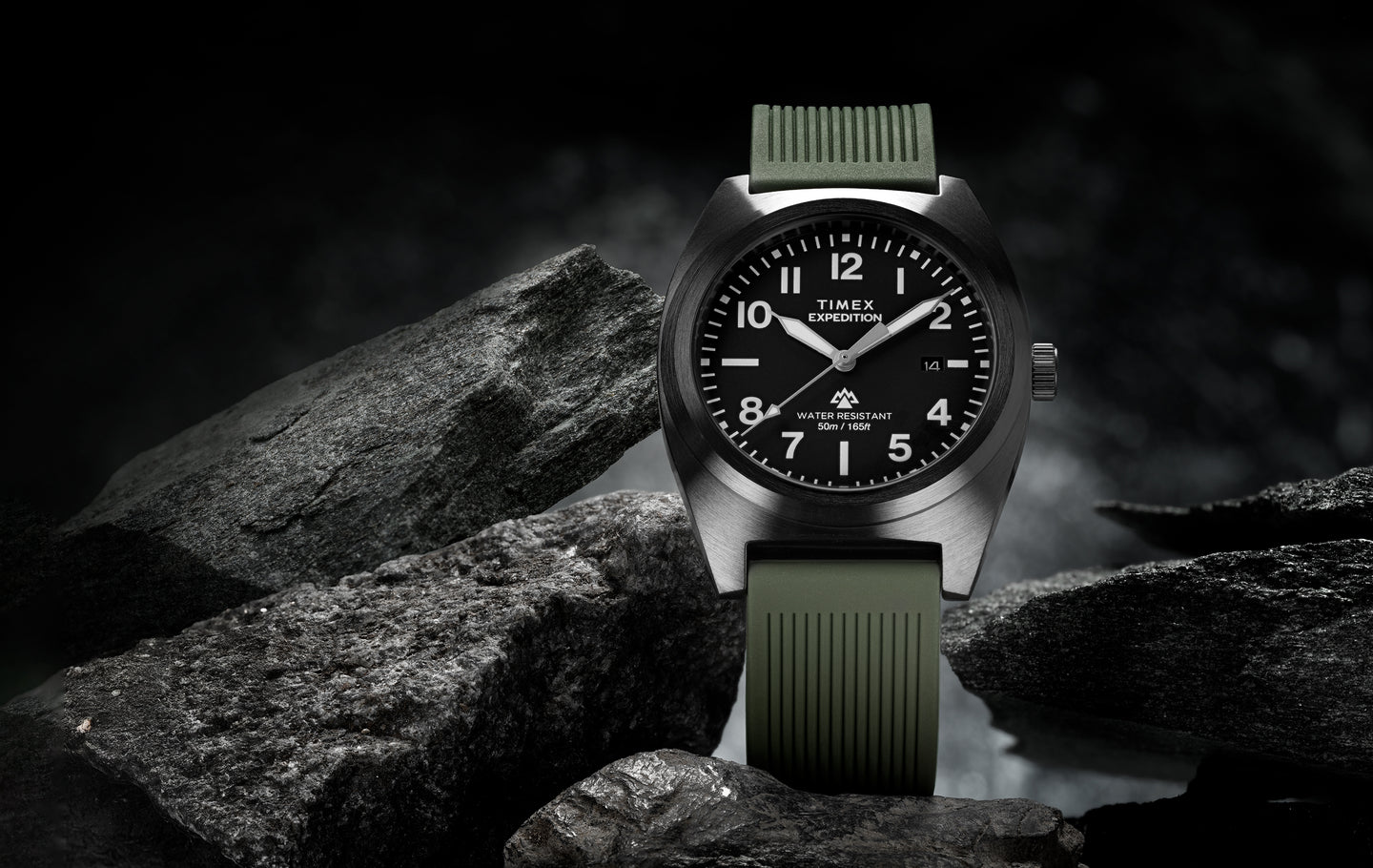A Timex Expedition® Capstone watch with a black dial, white numerals, and luminous hands, displayed among rugged gray rocks. The watch has a brushed stainless steel case, olive green textured strap, and is labeled “Water Resistant 50m / 165ft.” The dark, moody lighting emphasizes its durable, outdoor-ready design.