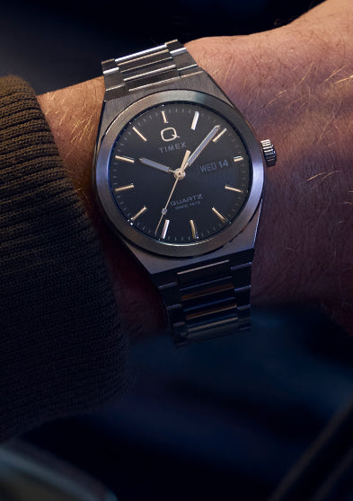 Wrist shot of the Q Timex® Continental Day and Date worn under a brown jacket sleeve. The stainless-steel bracelet and dark dial pair effortlessly for a timeless, understated style suited for both casual and formal wear.