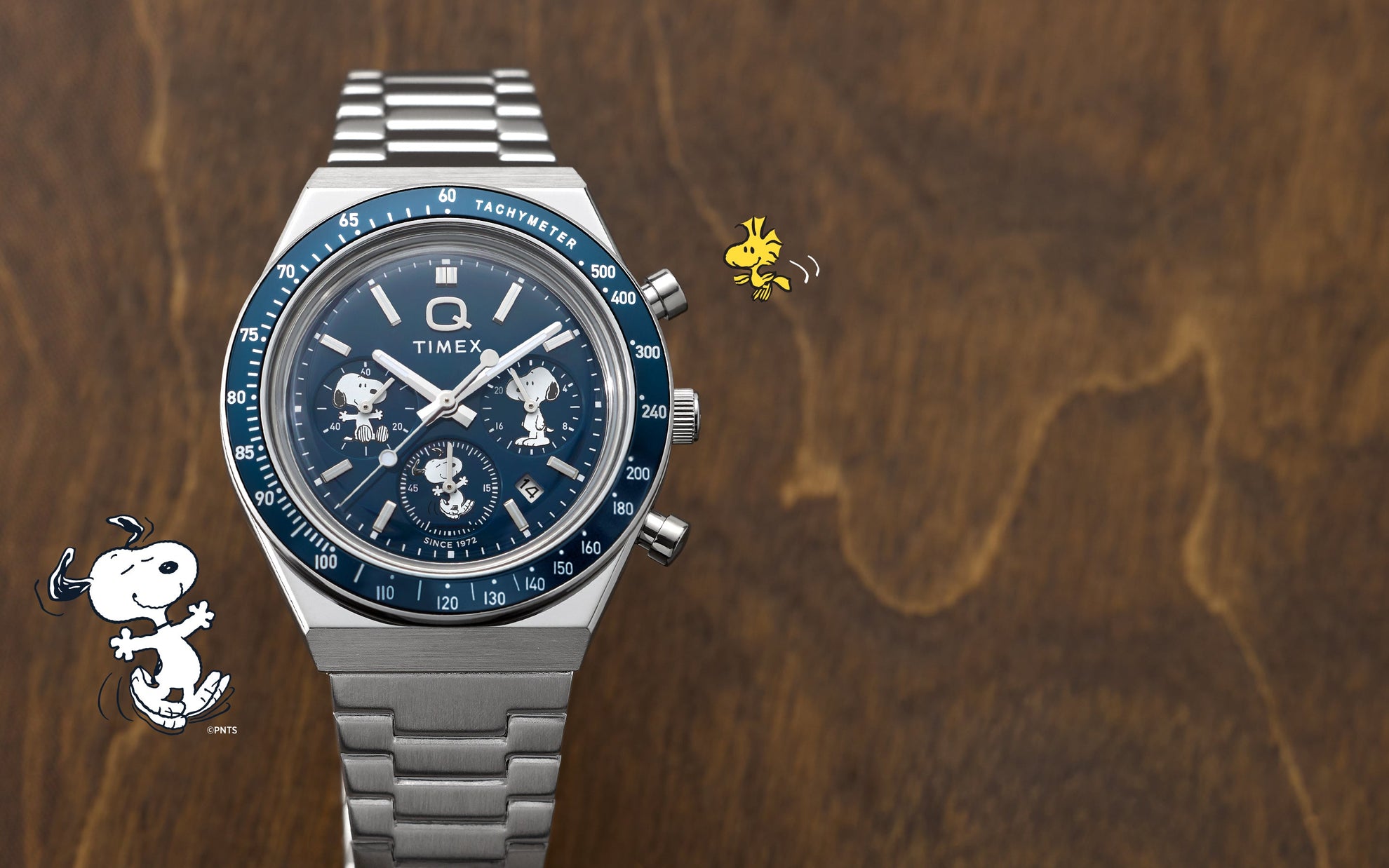 Timex x Peanuts® Q Timex® Chronograph resting on a wooden surface, showcasing its blue tachymeter bezel, silver-tone stainless steel bracelet, and Snoopy and Woodstock illustrations around the dial.