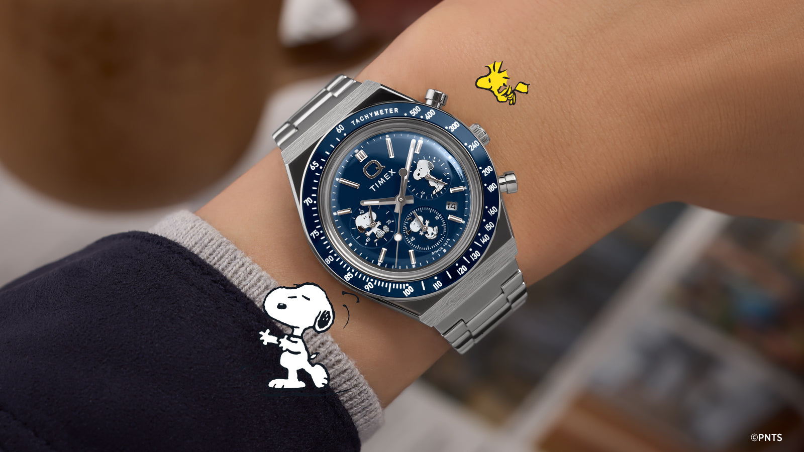 Stylized flat lay of the Timex x Peanuts® Q Timex® Chronograph with blue dial, silver-tone case, and Snoopy and Woodstock graphics, evoking a blend of playful nostalgia and timeless design.