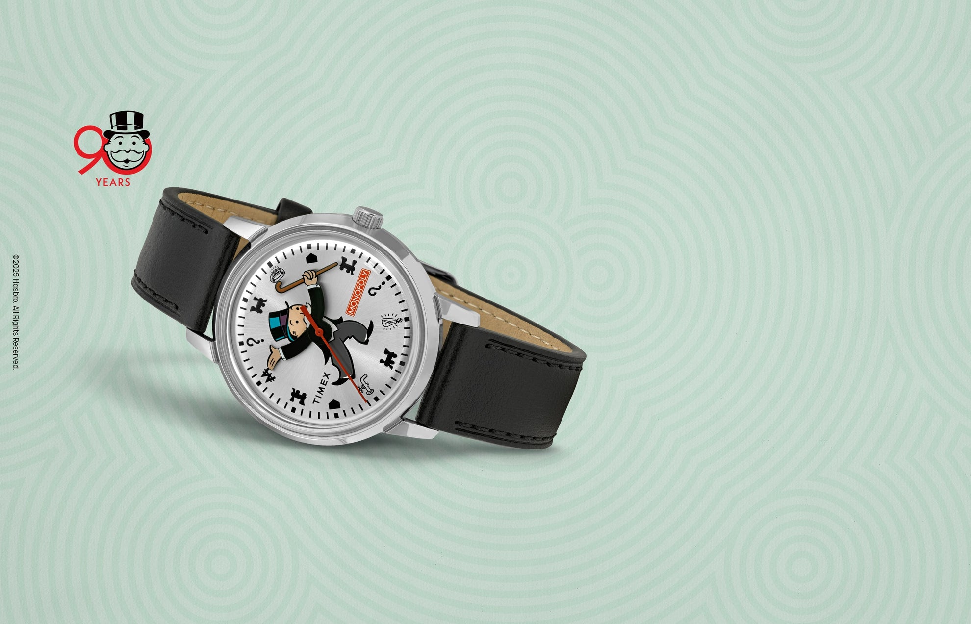 Timex x Monopoly Marlin® Automatic wristwatch with a silver sunburst dial featuring the Monopoly mascot Rich Uncle Pennybags as the central design. The Monopoly logo appears in red near the 3 o’clock position, surrounded by playful game icons including a top hat, dog, and question marks. The watch has a black leather strap and silver case, celebrating Monopoly’s 90th anniversary.