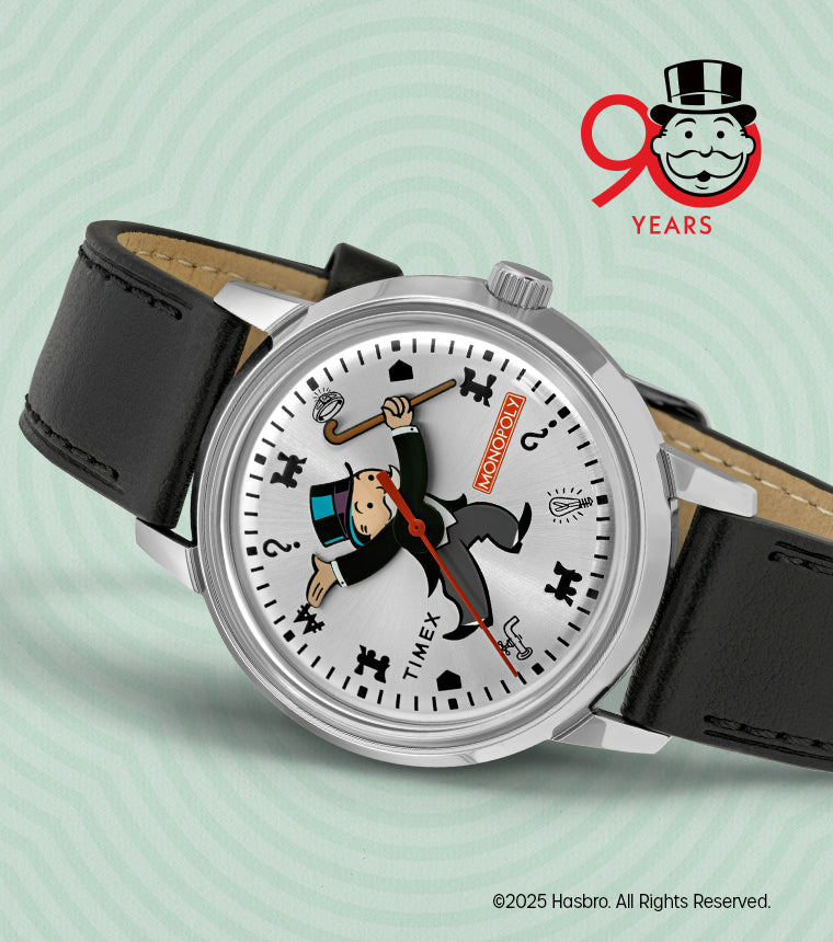 Timex x Monopoly Marlin® Automatic wristwatch with a silver sunburst dial featuring the Monopoly mascot Rich Uncle Pennybags as the central design. The Monopoly logo appears in red near the 3 o’clock position, surrounded by playful game icons including a top hat, dog, and question marks. The watch has a black leather strap and silver case, celebrating Monopoly’s 90th anniversary.