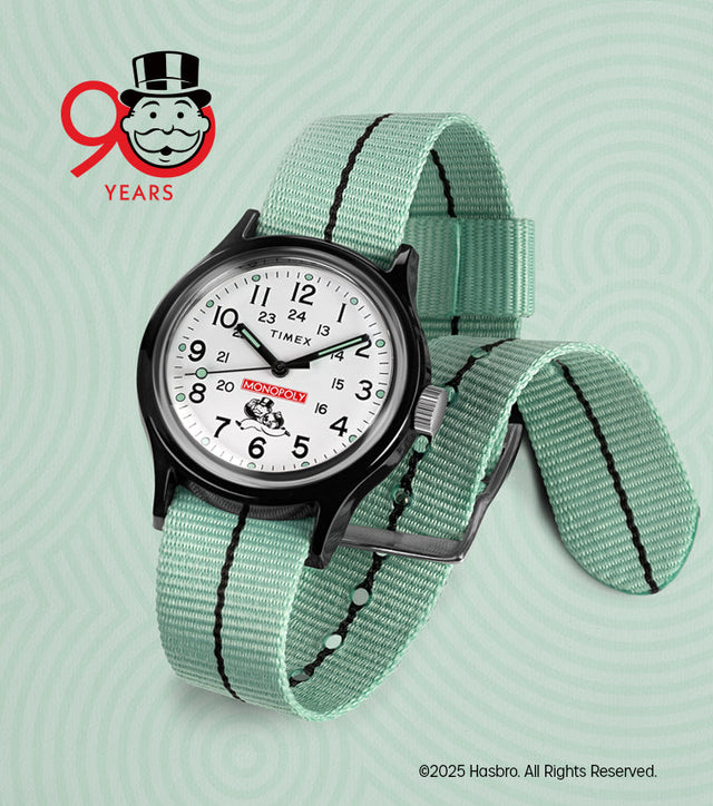A Timex x Monopoly MK1 wristwatch featuring a crisp white dial with black numerals, a red Monopoly logo above the 6 o’clock position, and the Monopoly mascot holding a money bag. The watch has a black case and a mint-green nylon strap. A “90 Years” Monopoly anniversary logo appears in the background.