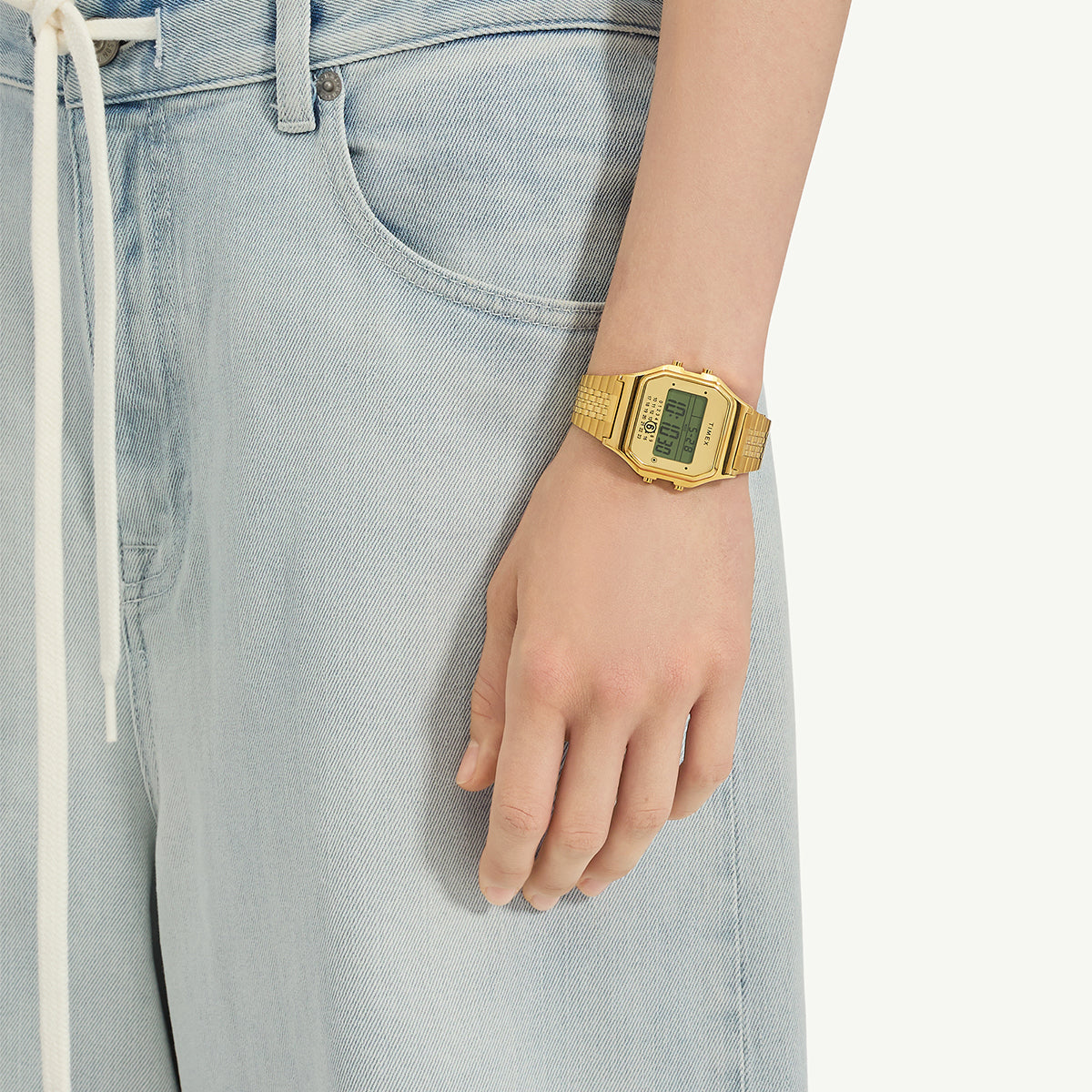 The gold-tone Timex x MM6 T80 watch worn on a wrist, paired with light blue denim jeans, showing its retro digital aesthetic and comfortable bracelet fit.