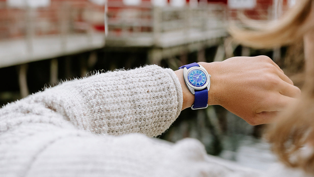 The Timex x Worn & Wound Whirlpool watch worn on a wrist outdoors, with a blue dial and strap visible against a waterfront setting and blurred red buildings in the background.