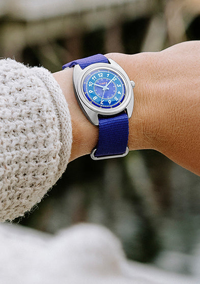 The Timex x Worn & Wound Whirlpool watch worn on a wrist outdoors, with a blue dial and strap visible against a waterfront setting and blurred red buildings in the background.