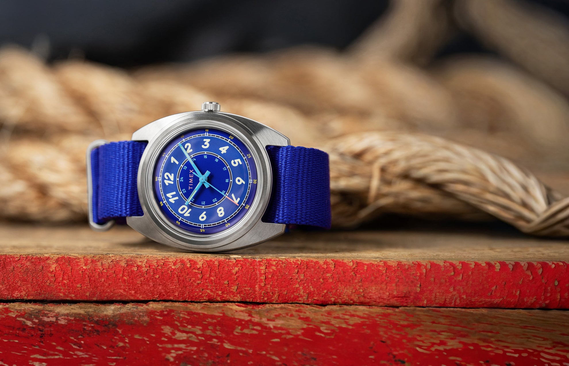 A Timex x Worn & Wound Whirlpool watch with a bright blue dial and matching blue fabric strap resting on a distressed red wooden surface, with thick coiled rope blurred in the background.