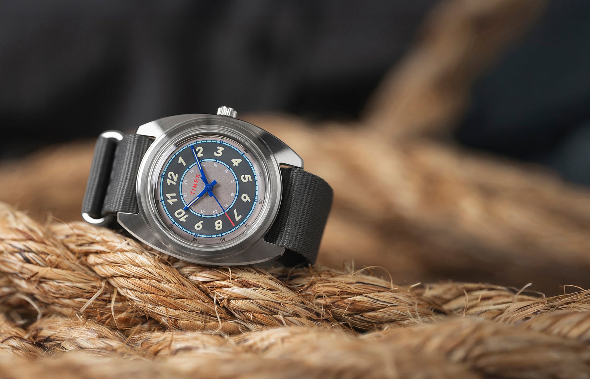A Timex x Worn & Wound Storm watch with a gray, blue, and silver multi-layered dial resting on thick coiled rope. The watch features cream numerals, blue hands, and a gray fabric strap.