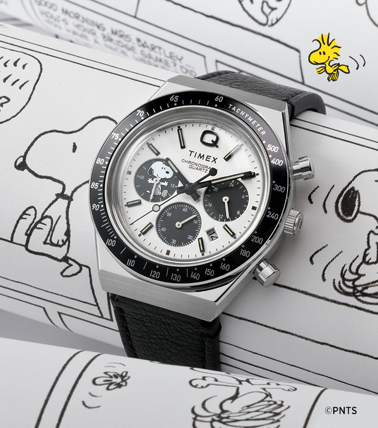 A Q Timex Chronograph x Peanuts Snoopy watch with a white dial, black sub-dials, and a black tachymeter bezel resting on Peanuts comic strips. The dial features Snoopy in one of the chronograph sub-dials, blending playful illustration with vintage racing-inspired design.