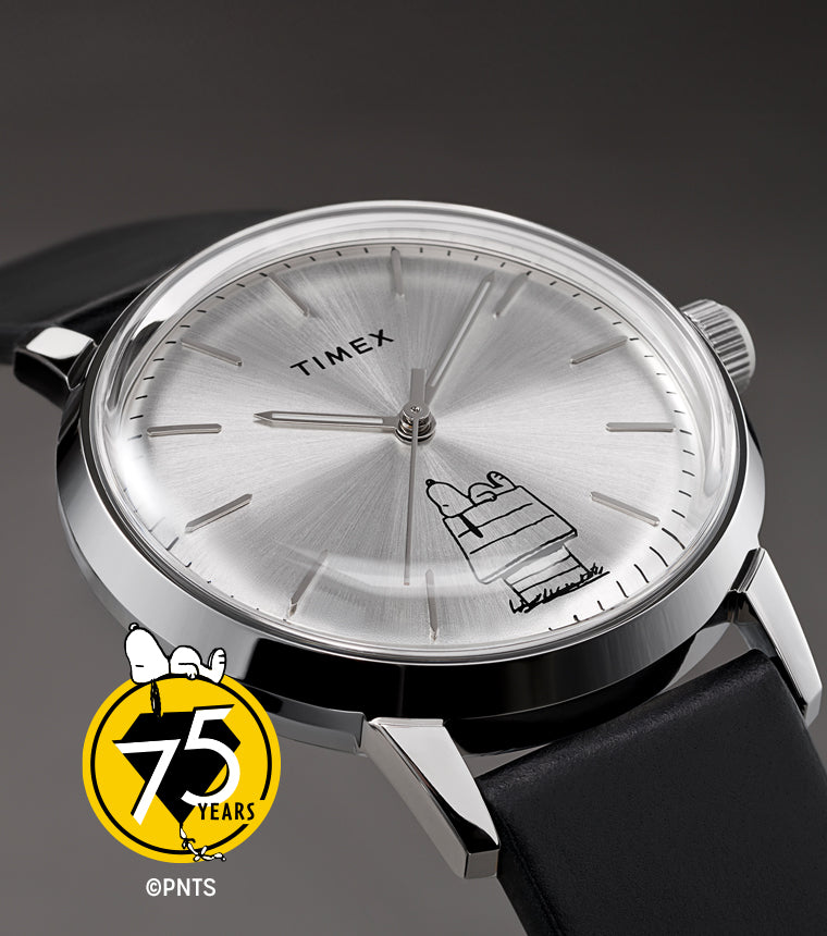 A Timex x Peanuts® Marlin® Automatic 75th Anniversary Edition watch with a silver sunburst dial and black leather strap displayed on a gray background. The dial features Snoopy lounging atop his doghouse in minimalist black line art, paired with the Peanuts 75th anniversary logo.