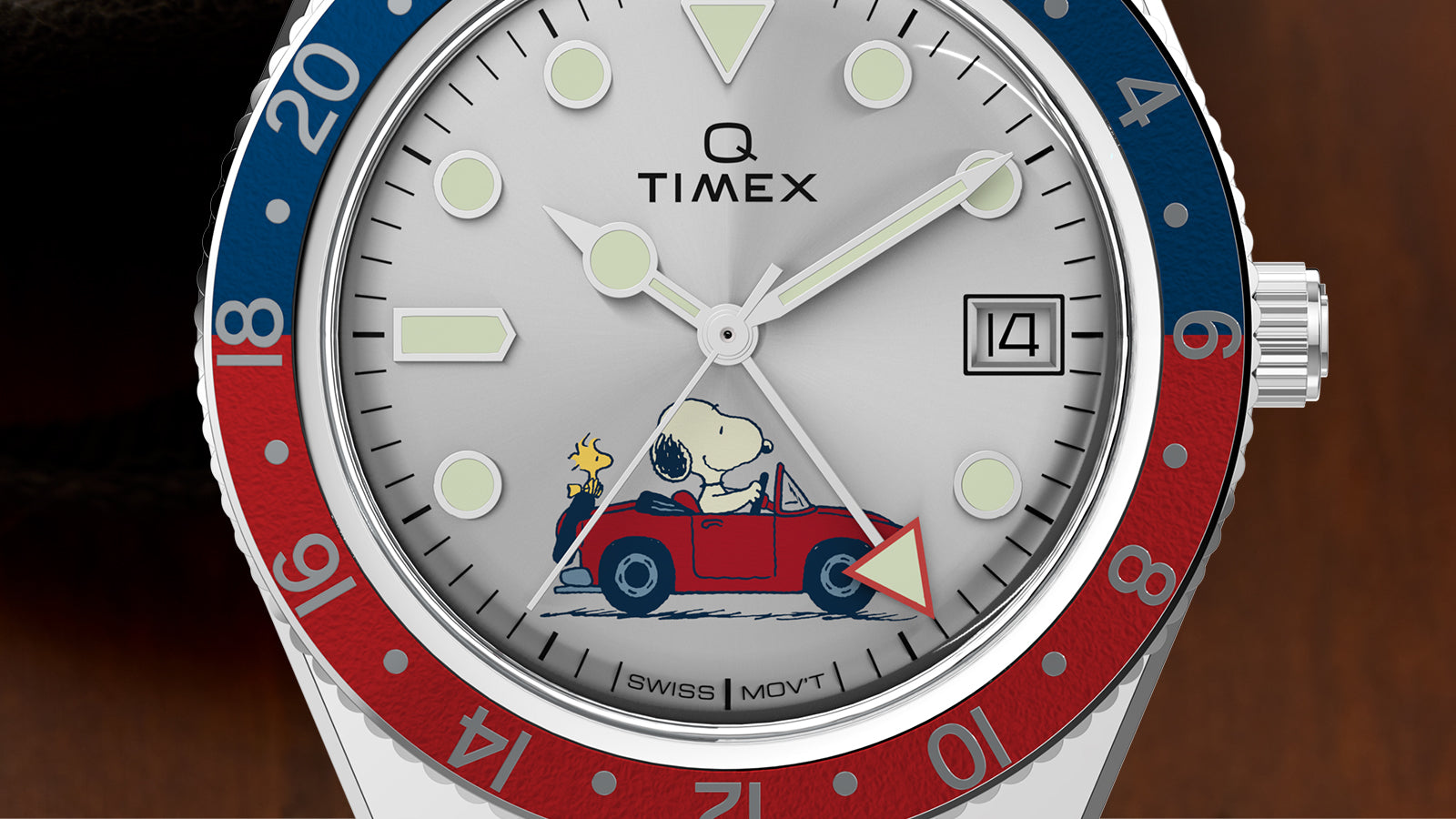 Close-up view of the Q Timex® GMT x Peanuts® watch dial showcasing Snoopy driving a red convertible with Woodstock beside him. The silver dial includes luminous hour markers, a red GMT hand, and the Timex logo beneath a domed crystal.