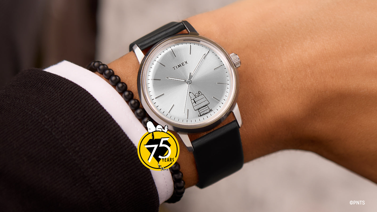 The Timex x Peanuts® Marlin® Automatic 75th Anniversary Edition watch worn on a wrist, highlighting its refined silver dial with Snoopy resting on his doghouse illustration and minimalist hour markers. The yellow Peanuts 75 Years logo adds a celebratory touch.