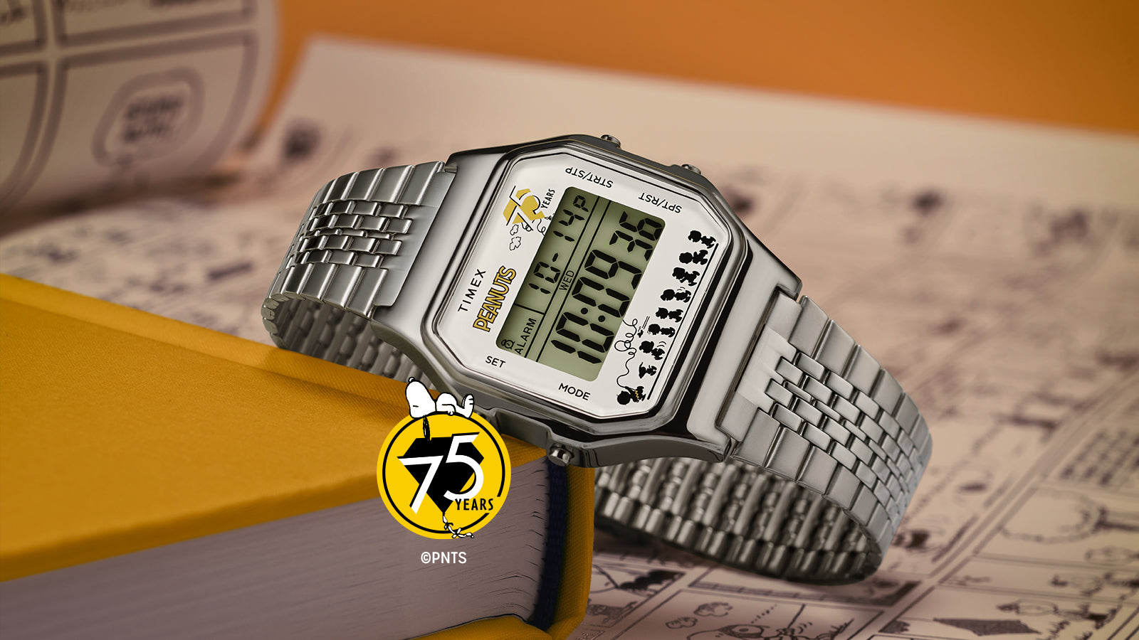 A Timex x Peanuts® T80 75th Anniversary Edition digital watch with a silver-tone stainless-steel bracelet resting on a yellow book. The display features the Peanuts 75 Years logo and silhouettes of the Peanuts characters walking along the bottom edge, set against a background of comic strip pages.
